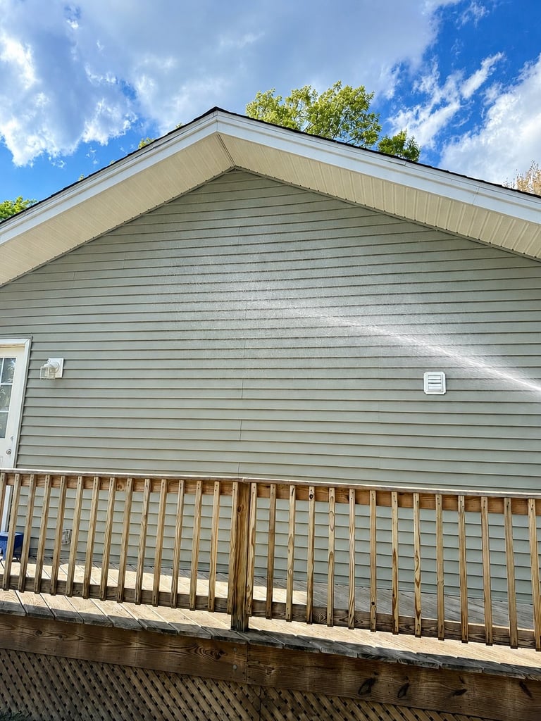 Gray sided house with white trim, gable roof, and wooden deck railing under blue sky with white clouds