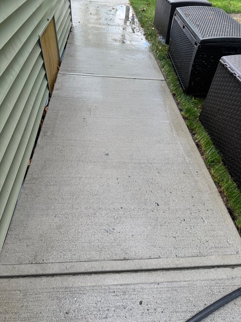 Narrow concrete walkway between buildings with wet surface, metal storage boxes, and grass edges
